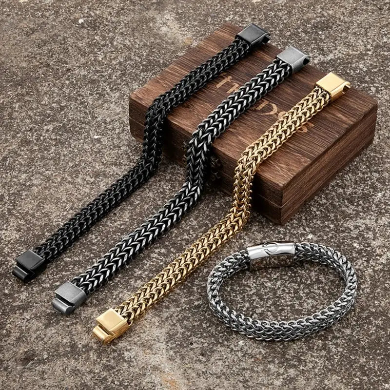 Three braided metal bracelets in black, silver, and gold tones are arranged on a textured stone surface with a wooden box. One bracelet is laid flat, while the other two are coiled, showing their chain links and clasp details.