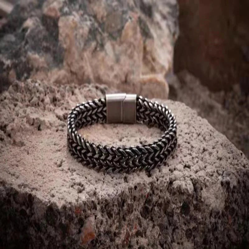 A braided metal bracelet with a silver-toned rectangular clasp, resting on a textured stone surface with a blurred natural rock background.