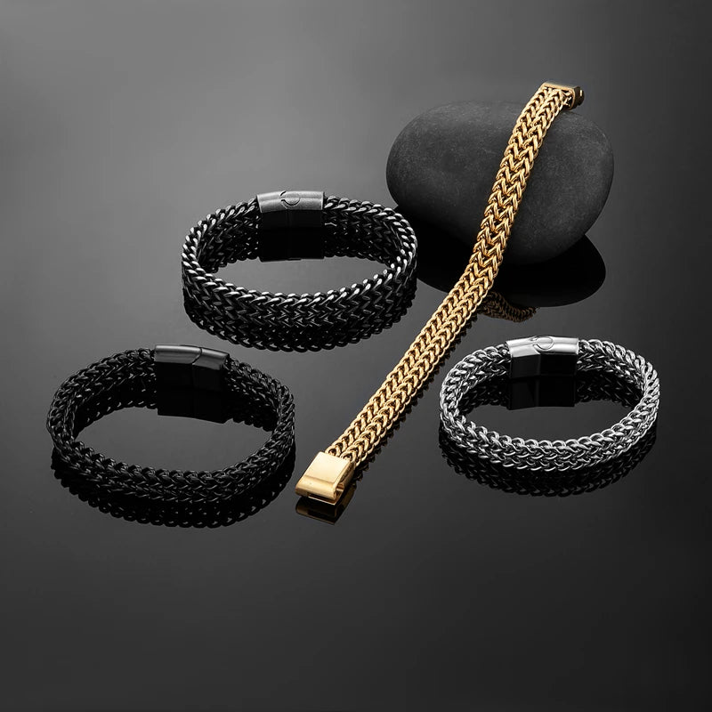 Four braided chain bracelets arranged on a glossy black reflective surface, with one gold-toned bracelet draped over a dark stone. The bracelets are black, silver, and gold-toned, each with a dark metal clasp.