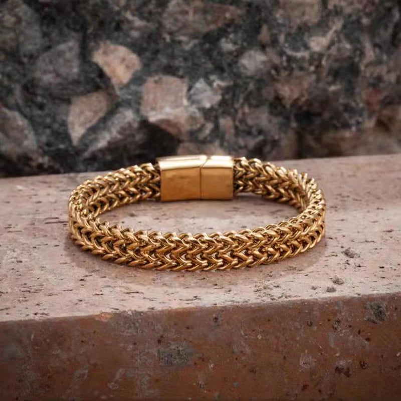 A gold-toned braided chain bracelet with a rectangular clasp, resting on a textured stone surface with a blurred rock background.