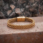 A gold-toned braided chain bracelet with a rectangular clasp, resting on a textured stone surface with a blurred rock background.