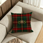 A square throw pillow with a green and red tartan plaid pattern and a central holly and snowflake motif, resting on a light beige fabric sofa in a living room setting.