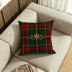 A square throw pillow with a green and red tartan plaid pattern and a central holly and snowflake motif, resting on a light beige fabric sofa in a living room setting.