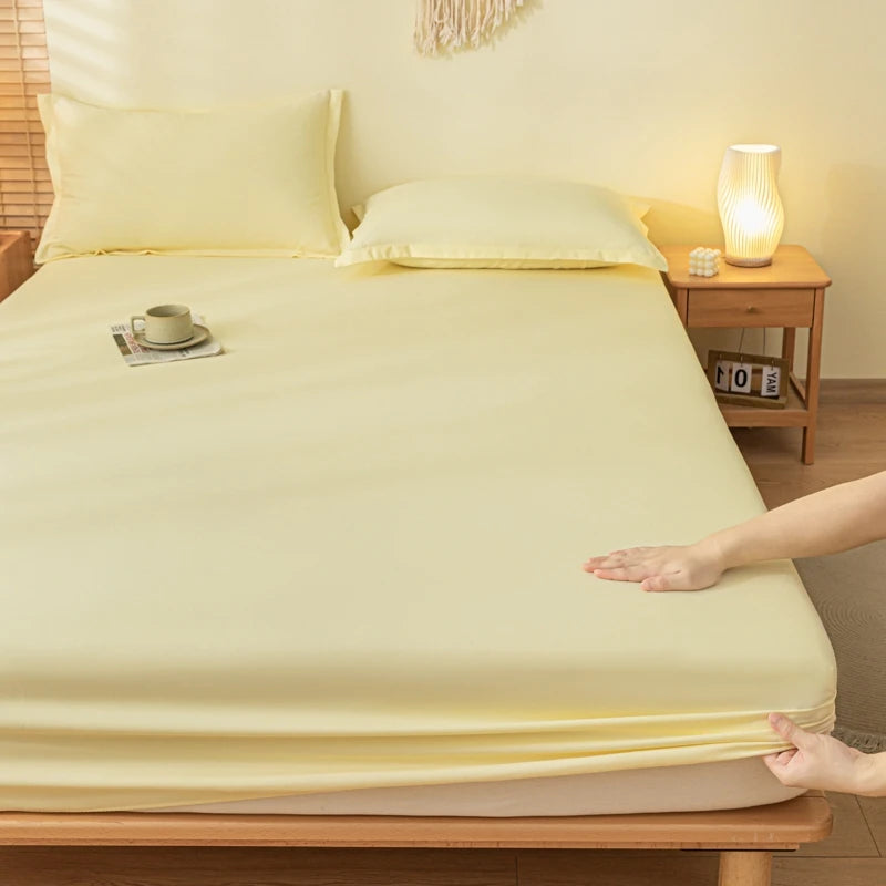 A pale yellow fitted sheet and matching pillowcases on a bed in a warm, softly lit bedroom. A person's hands are shown pulling the sheet tightly at the corner, demonstrating its stretch.