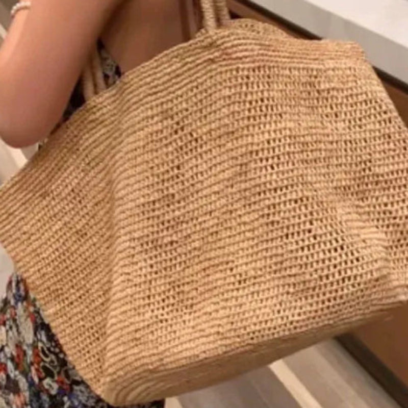 A woman in a floral dress carries a large woven straw tote bag with double shoulder straps in a modern indoor setting with wood cabinetry and a marble countertop.
