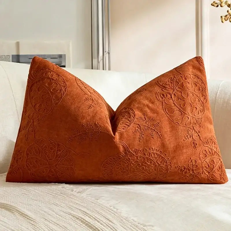 An orange-brown decorative throw pillow with an embossed floral pattern, resting on a cream-colored sofa.