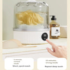 A countertop fabric cleaner or steamer with a clear dome lid, white base, and digital control panel, shown with a hand pressing the button. A yellow cloth is inside, and a soap dispenser sits beside it.