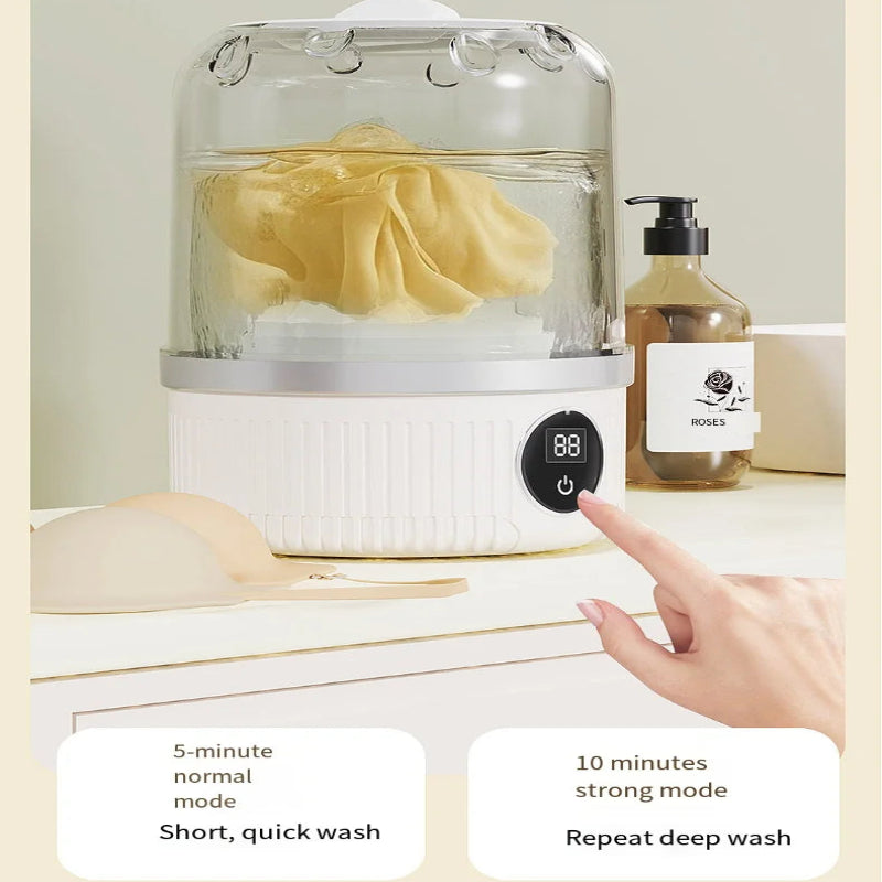 A countertop fabric cleaner or steamer with a clear dome lid, white base, and digital control panel, shown with a hand pressing the button. A yellow cloth is inside, and a soap dispenser sits beside it.