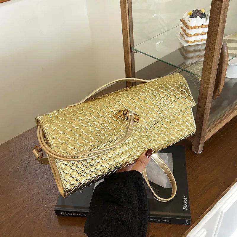 Gold woven handbag held by a person on a wooden surface with a glass cabinet in the background.