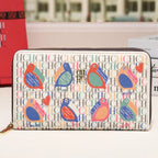 A long zip-around wallet with a colorful bird and heart pattern over a repeating monogram background, displayed on a white surface with a blurred building facade in the background.
