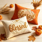 Two decorative throw pillows with embroidered pumpkins and the word 'blessed' in autumn colors, styled on a light wood surface with small pumpkins, wheat, and fall leaves.