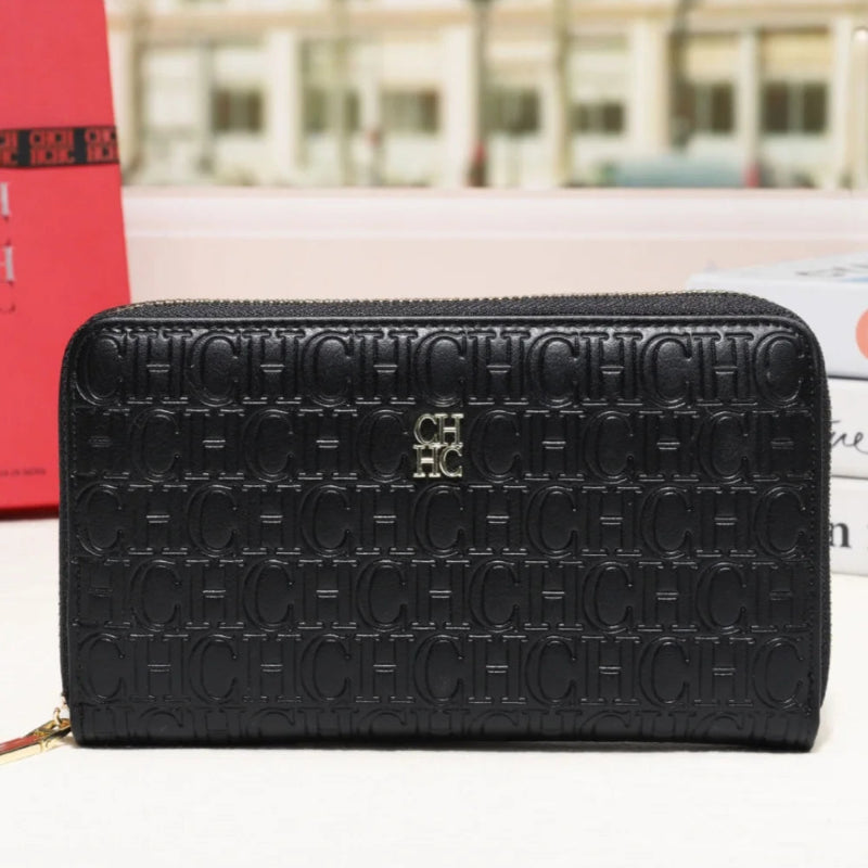 A black leather zip-around wallet with an embossed monogram pattern and a small gold-tone logo at the center front, displayed on a white surface with a blurred city street background.