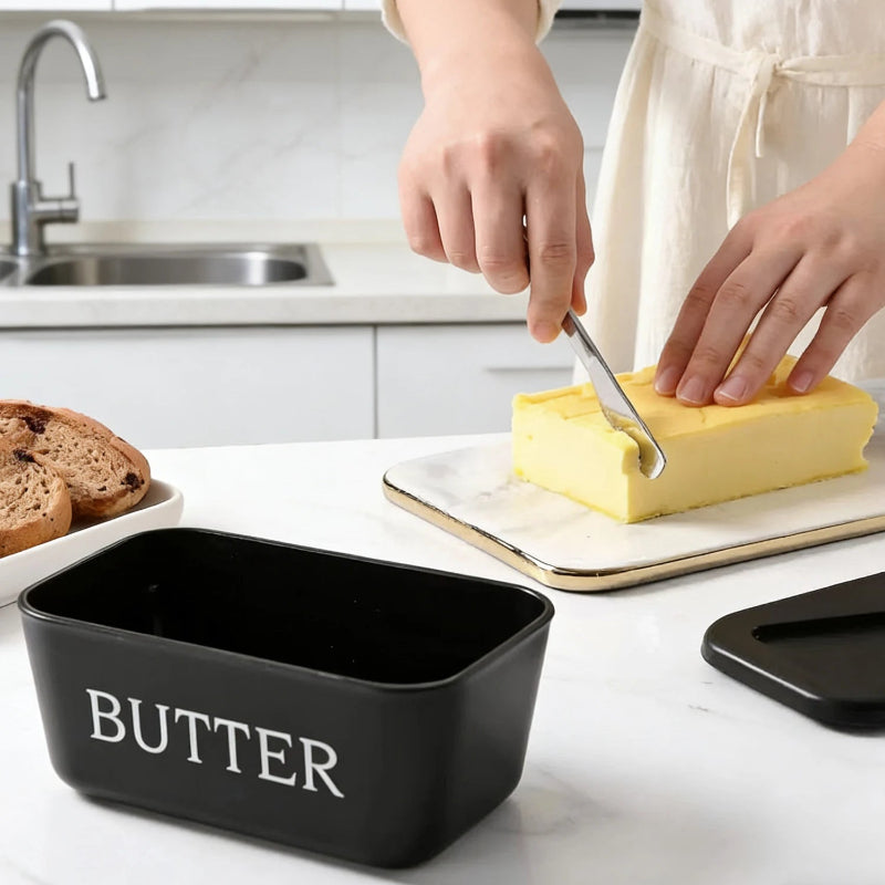 Person cutting butter with a knife on a kitchen counter next to a black butter container.