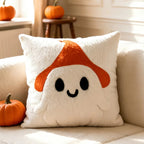 A white textured throw pillow featuring a cute ghost character with an orange mushroom cap and black embroidered eyes and smile, resting on a beige sofa with pumpkins nearby.