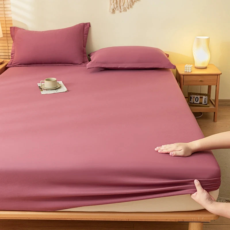 A fitted sheet and matching pillowcases in a dusty rose or mauve color are shown on a bed in a styled bedroom. A person's hands are visible at the bottom right pulling the sheet tightly, demonstrating its stretch.