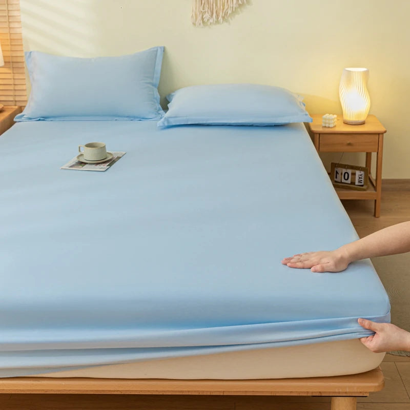 A light blue fitted sheet and matching pillowcases on a bed, with a person's hands pulling the sheet down over a mattress. A wooden nightstand with a glowing lamp is visible in the background of the bedroom setting.