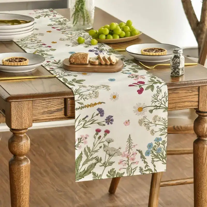 A floral table runner with a cream background and multicolored wildflower print is styled on a wooden dining table with plates, bread, green grapes, and a pepper shaker.