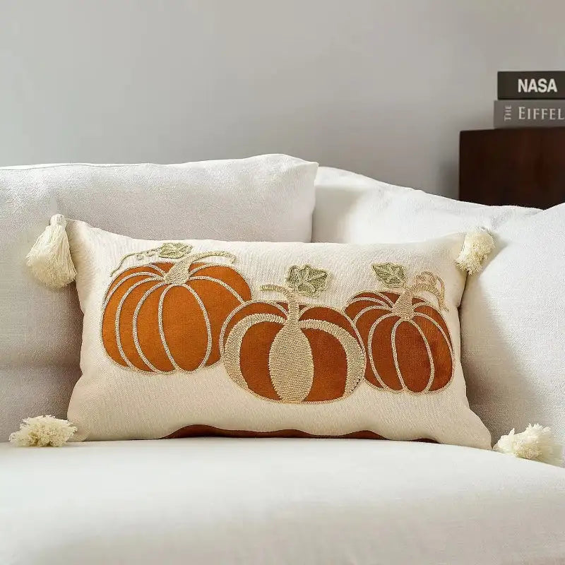 A rectangular cream throw pillow with embroidered orange pumpkins and tassels, styled on a white sofa with a book stack in the background.