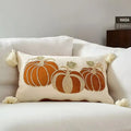 A rectangular cream throw pillow with embroidered orange pumpkins and tassels, styled on a white sofa with a book stack in the background.