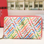 A colorful zip-around wallet with a repeating monogram pattern in red, blue, green, yellow, and pink on a white base, shown on a tabletop with a blurred building background.
