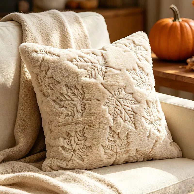 A square cream throw pillow with raised leaf embroidery, styled on a beige sofa next to a matching blanket, with a pumpkin in the background.
