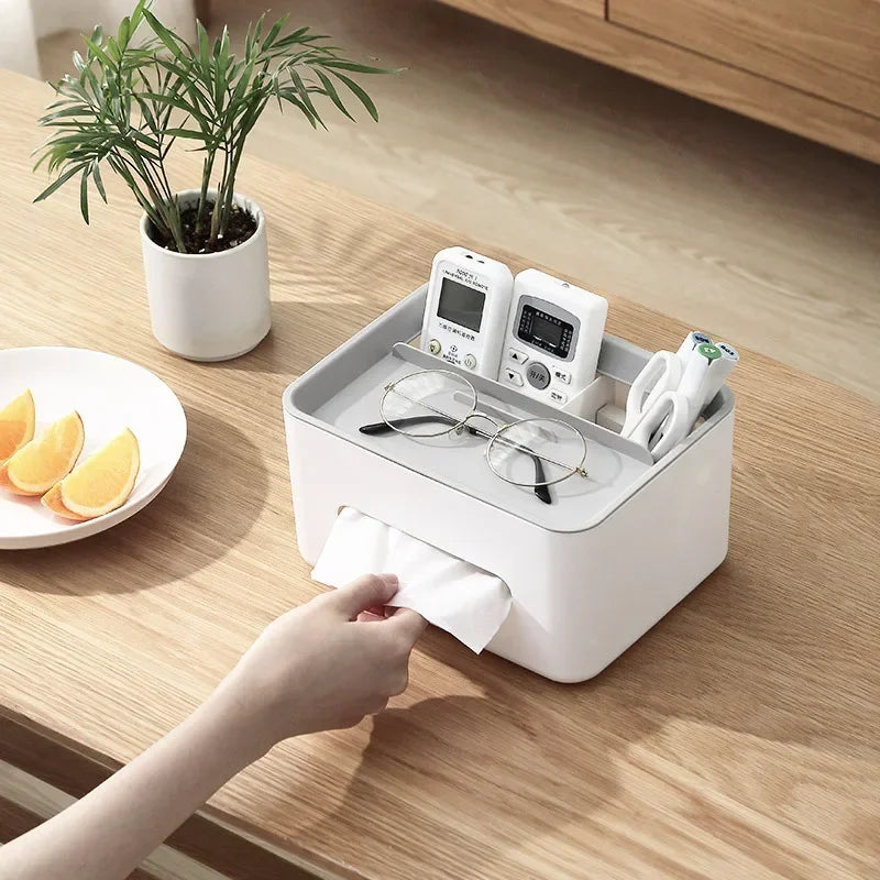 White tissue box on a wooden table with a hand reaching for a tissue, next to a plate of fruit and a plant.