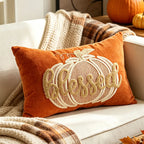 An orange velvet throw pillow with a large embroidered pumpkin and the word 'Blessed' in cream, styled on a white couch with autumn blankets and pumpkins in the background.