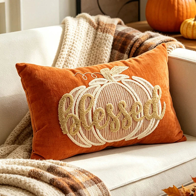 An orange velvet throw pillow with a large embroidered pumpkin and the word 'Blessed' in cream, styled on a white couch with autumn blankets and pumpkins in the background.