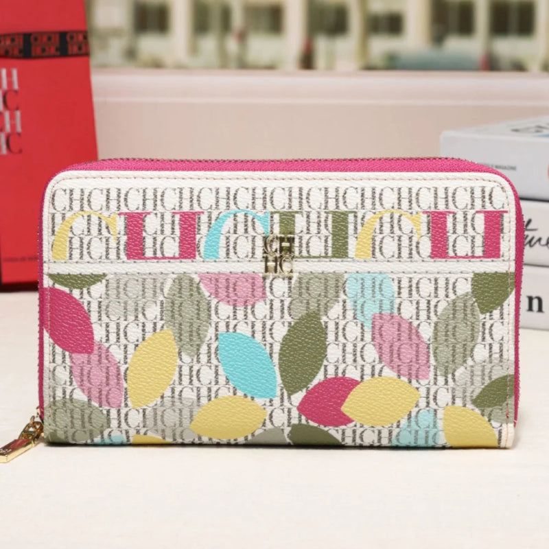 A zip-around wallet with a pink border and a colorful leaf pattern over a white monogram background, displayed on a tabletop with a blurred building facade in the background.