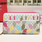 A zip-around wallet with a pink border and a colorful leaf pattern over a white monogram background, displayed on a tabletop with a blurred building facade in the background.