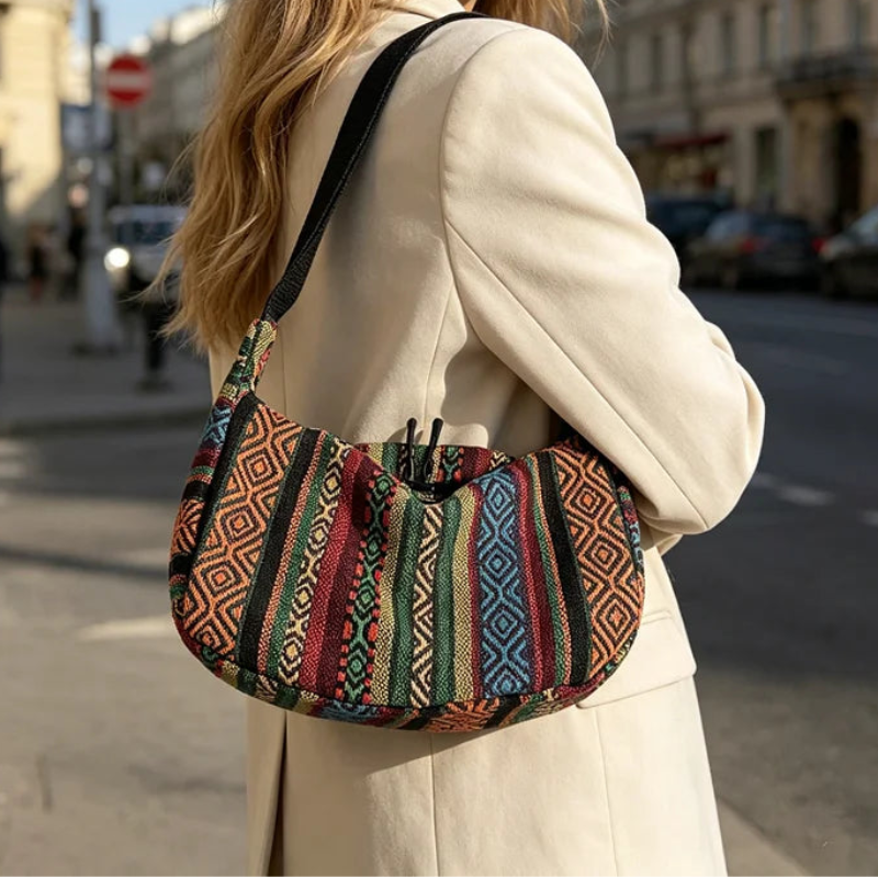 A woman in a cream coat carries a slouchy shoulder bag with a colorful geometric tribal pattern in red, blue, green, and brown, set against a blurred city street background.