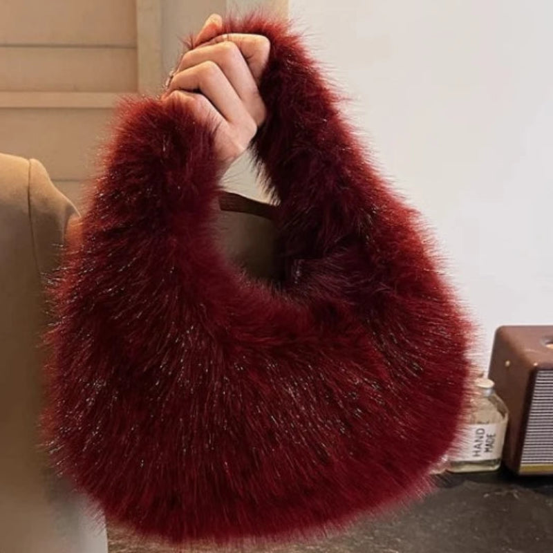 A person in a beige blazer holds up a large, fuzzy crescent-shaped shoulder bag in deep burgundy red. The background shows a neutral indoor setting with a wooden slatted door and a vintage-style speaker on a table.