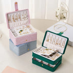 Three velvet jewelry boxes in pink, blue, and green, shown open to display their interior compartments filled with earrings and jewelry. The boxes are arranged on a light beige tabletop with a soft, neutral background and a decorative lantern.