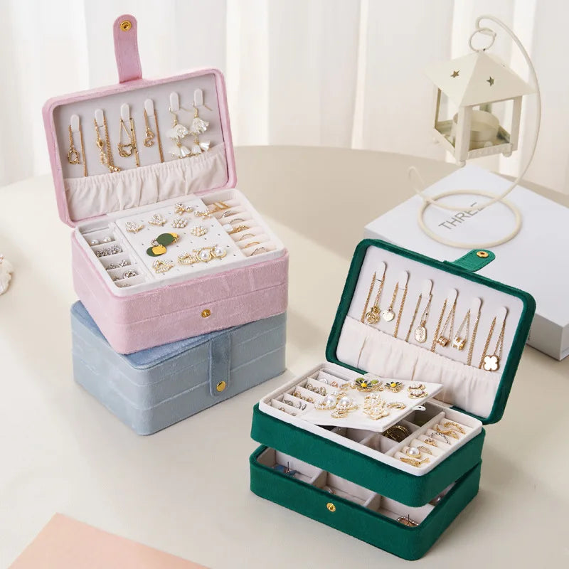 Three velvet jewelry boxes in pink, blue, and green, shown open to display their interior compartments filled with earrings and jewelry. The boxes are arranged on a light beige tabletop with a soft, neutral background and a decorative lantern.