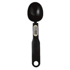 A digital kitchen measuring spoon in matte black plastic with an integrated LCD display showing '0.0' and a small beige accent ring, set against a plain white background.
