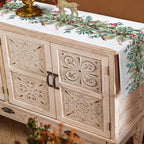 A white Christmas table runner with a green pine garland and red berry pattern, draped over a distressed white carved wooden sideboard cabinet in a warm brown room.