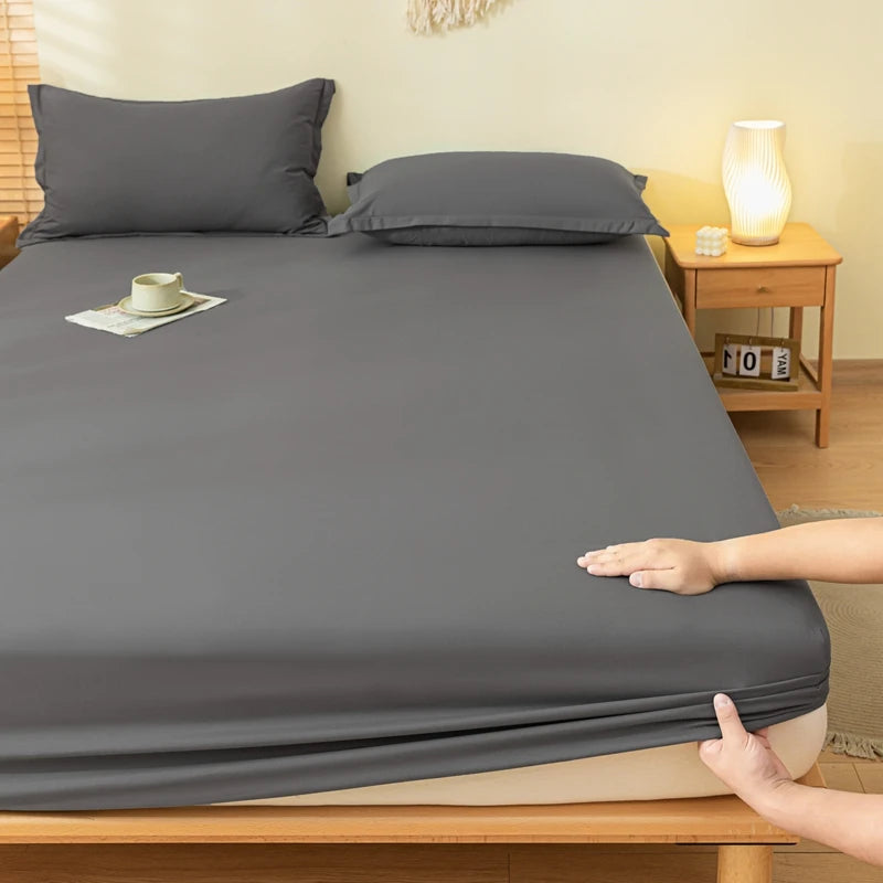 A dark gray fitted sheet and matching pillowcases on a bed, with a person's hands pulling the sheet up over the mattress corner in a bedroom setting.