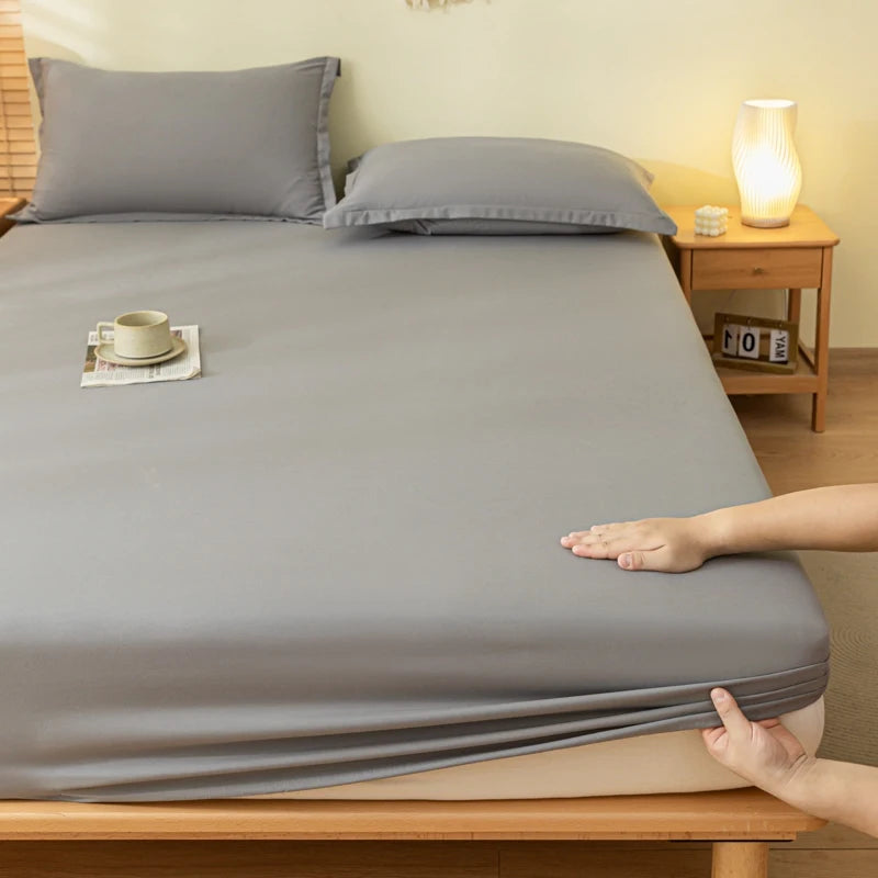 A gray fitted sheet and matching pillowcases on a wooden bed frame, with a person's hands pulling the sheet down over the mattress. A small side table with a lit lamp and a cup sits in the background.