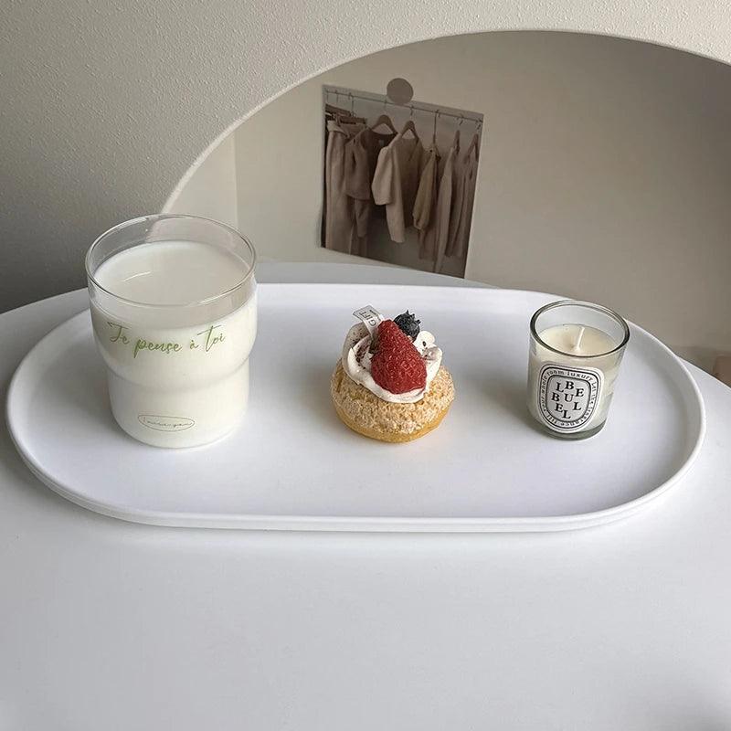 Tray with a glass of milk, a small cake, and a candle on a white surface.