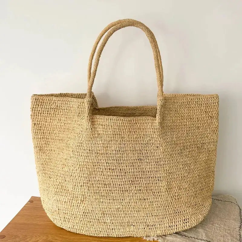 Natural woven straw tote bag with two top handles, displayed on a wooden surface against a plain off-white wall.