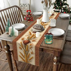 A rectangular woven table runner with a beige and brown holiday wheat design is styled on a wooden dining table. The table is set with white plates, candles, a white pitcher, and small decorative Christmas trees, with a Christmas tree visible in the background.