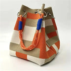 A canvas bucket bag with a geometric checkered pattern in orange, beige, and white, featuring orange braided rope handles with blue accents and silver metal hardware.