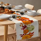 Dining table set with a floral and harvest-themed table runner, plates, bowls, and glasses.