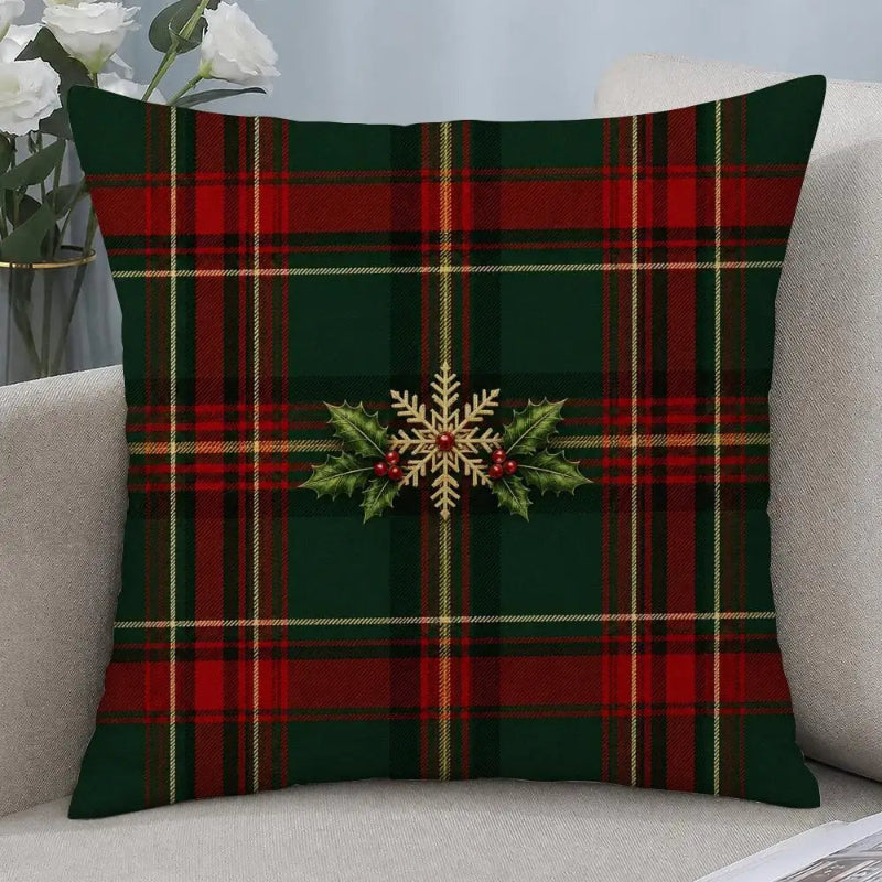 A square throw pillow with a red and green tartan plaid pattern and a central gold snowflake and holly design, displayed on a light beige sofa.