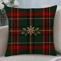 A square throw pillow with a red and green tartan plaid pattern and a central gold snowflake and holly design, displayed on a light beige sofa.