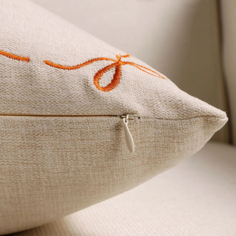 Close-up of a beige textured throw pillow with orange embroidered script and a silver zipper pull, resting on a neutral surface.