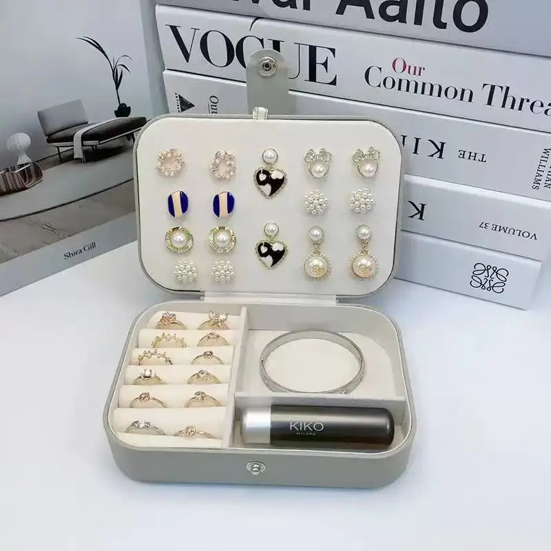 An open jewelry organizer case displaying earrings, rings, a bracelet, and a lip balm. The case is styled in front of stacked fashion books, including Vogue and The King of Thieves.