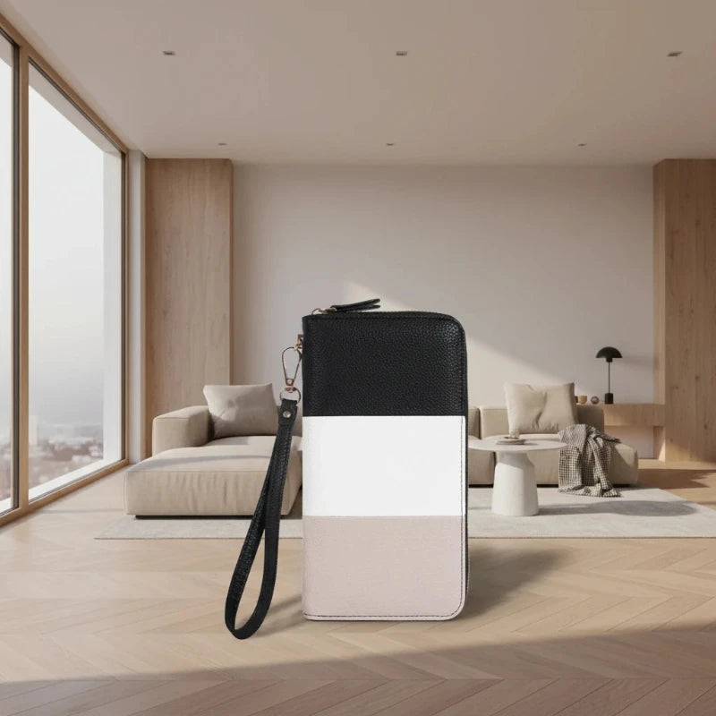 A color-blocked zippered wallet or clutch with a black, white, and light beige design, featuring a black wrist strap, set against a modern, sunlit minimalist living room background.