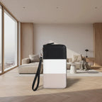 A color-blocked zippered wallet or clutch with a black, white, and light beige design, featuring a black wrist strap, set against a modern, sunlit minimalist living room background.