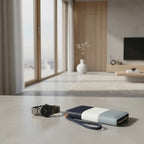 A color-blocked leather wallet with a wrist strap lies on a light stone surface in a modern, sunlit living room. The wallet features navy blue, white, and light gray sections.